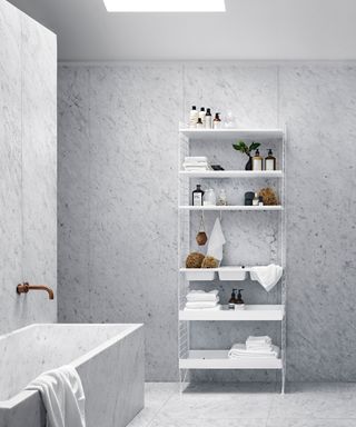 Minimalist, modern bathroom with string shelving white unit, decorated with towels, toiletries, decorative objects, matching marble tiles on walls and floors, matching tiled bath with copper tap