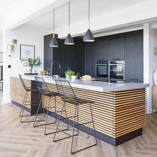 Large kitchen island clad in wooden batons with tall black units behind