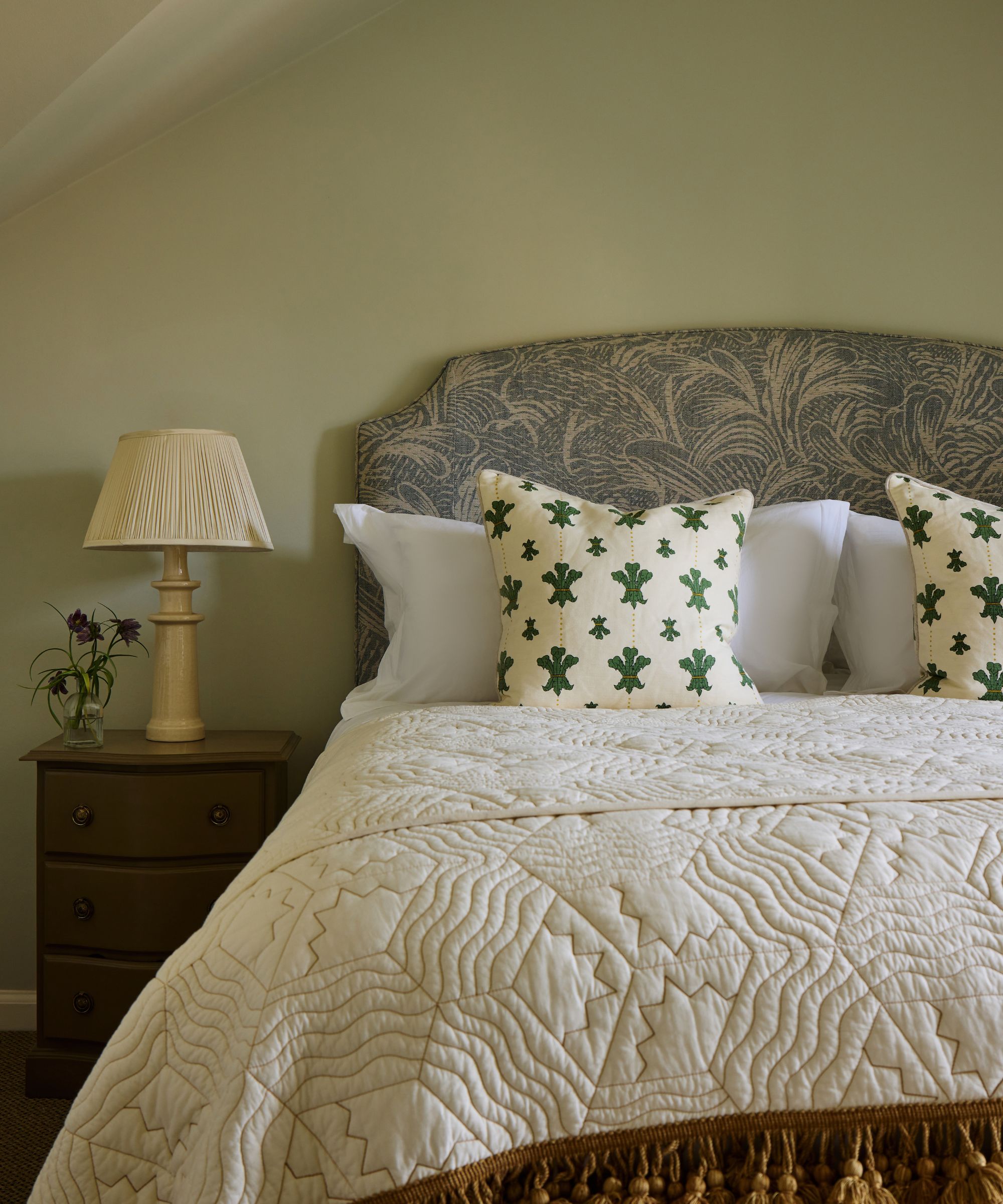 A cozy bed with a gray patterned headboard, quilted cream coverlet, and green-patterned throw pillows, next to a wood nightstand with a classic lamp against soft sage walls