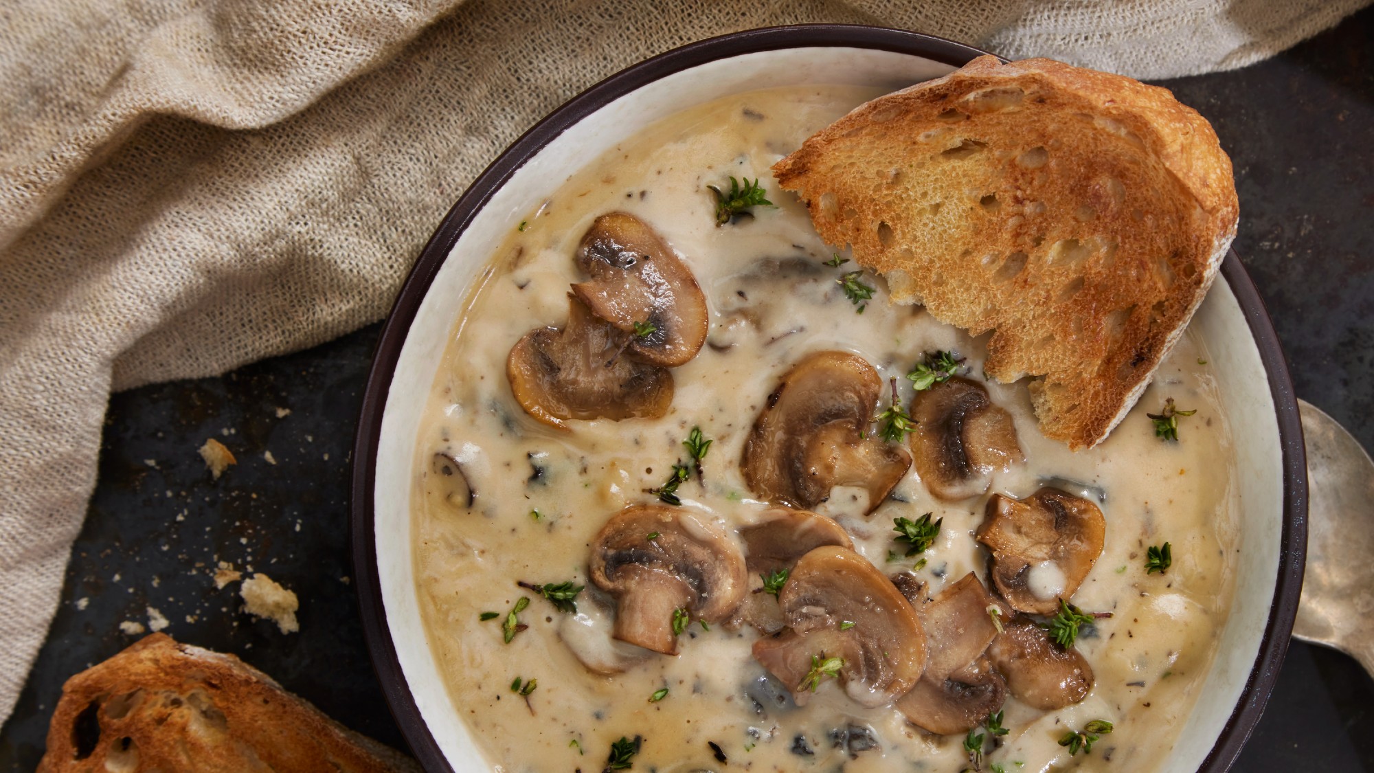 overhead shot of a bowl of mushroom soup. a large slice of toasted bread is set atop the bowl. a linen napkin is draped across the left top of the photo
