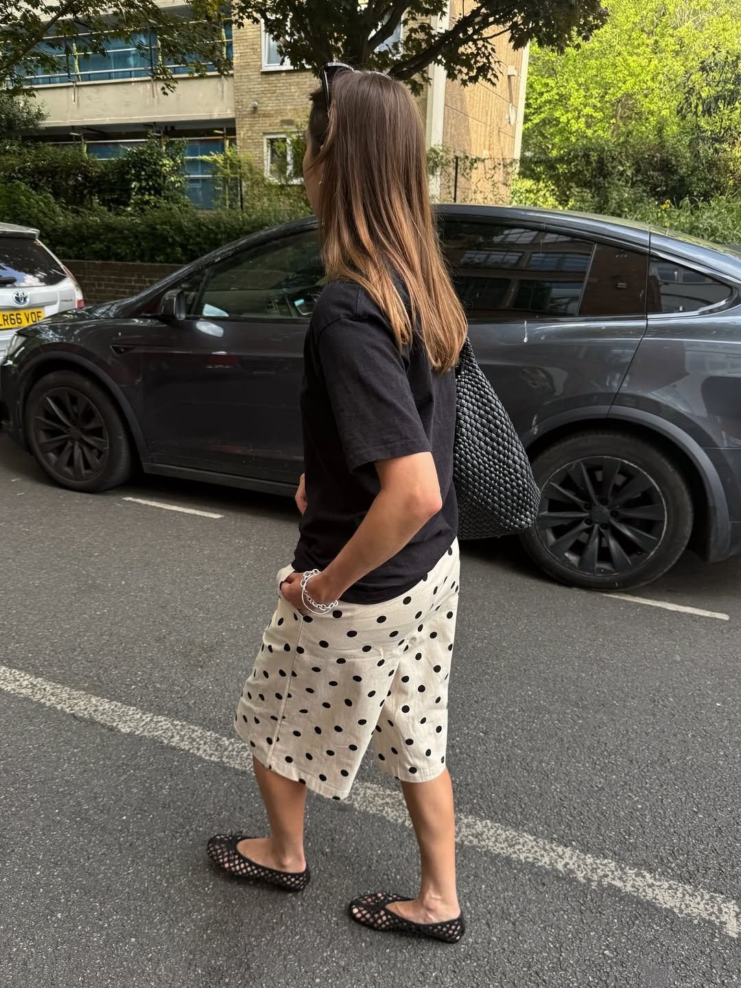 Influencer Poppy wears polka dot long shorts with a black crewneck top, a woven bag, and mesh ballet flats.