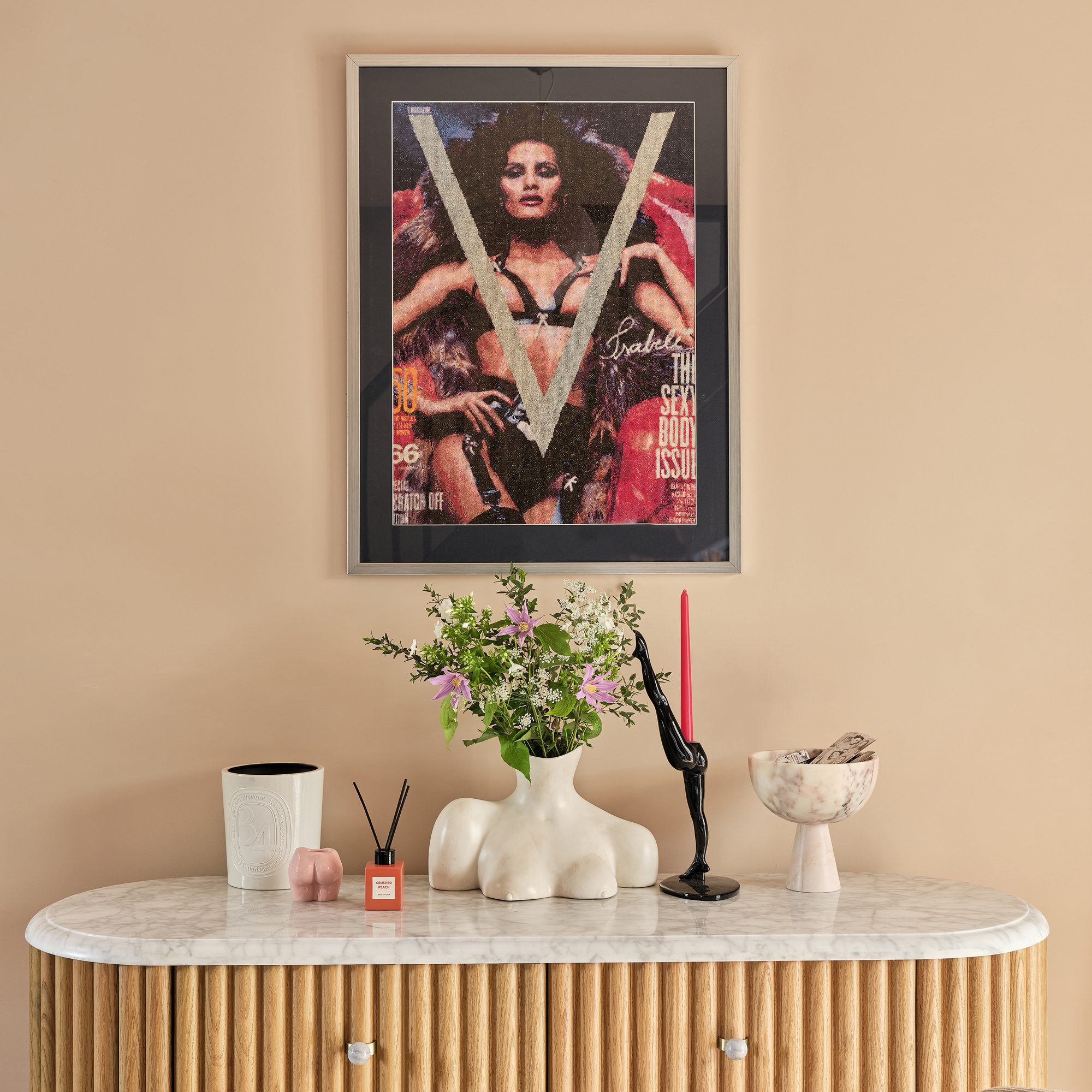 a fluted wood chest of drawers with a marble top with a selection of accessories and vases and a framed magazine cover on the wall