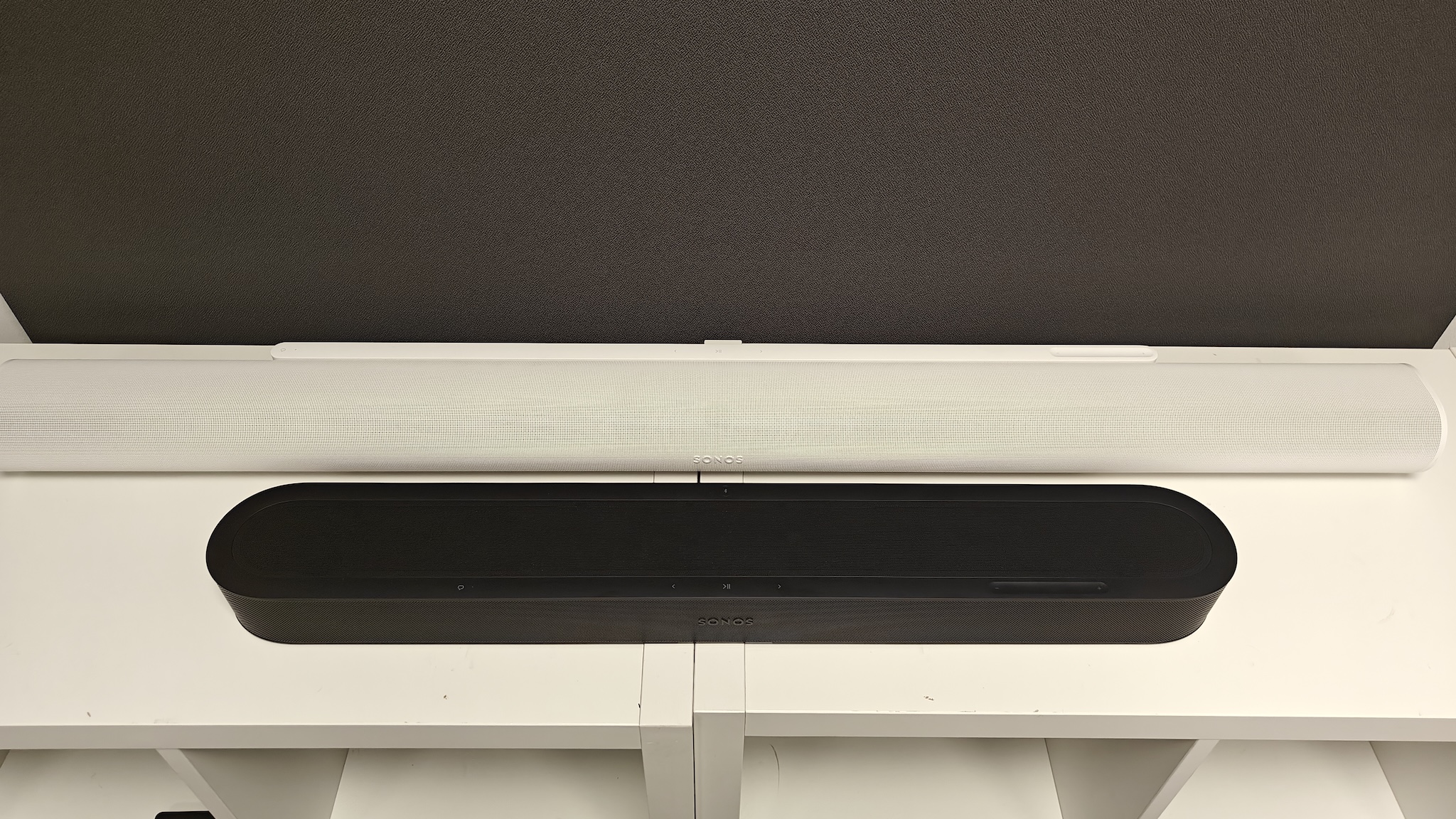 A black Sonos Beam Ultra and white Sonos Arc Ultra next to each other on a white TV cabinet, shot from above and cropped slightly tight so that one end of the Arc Ultra is out of frame.