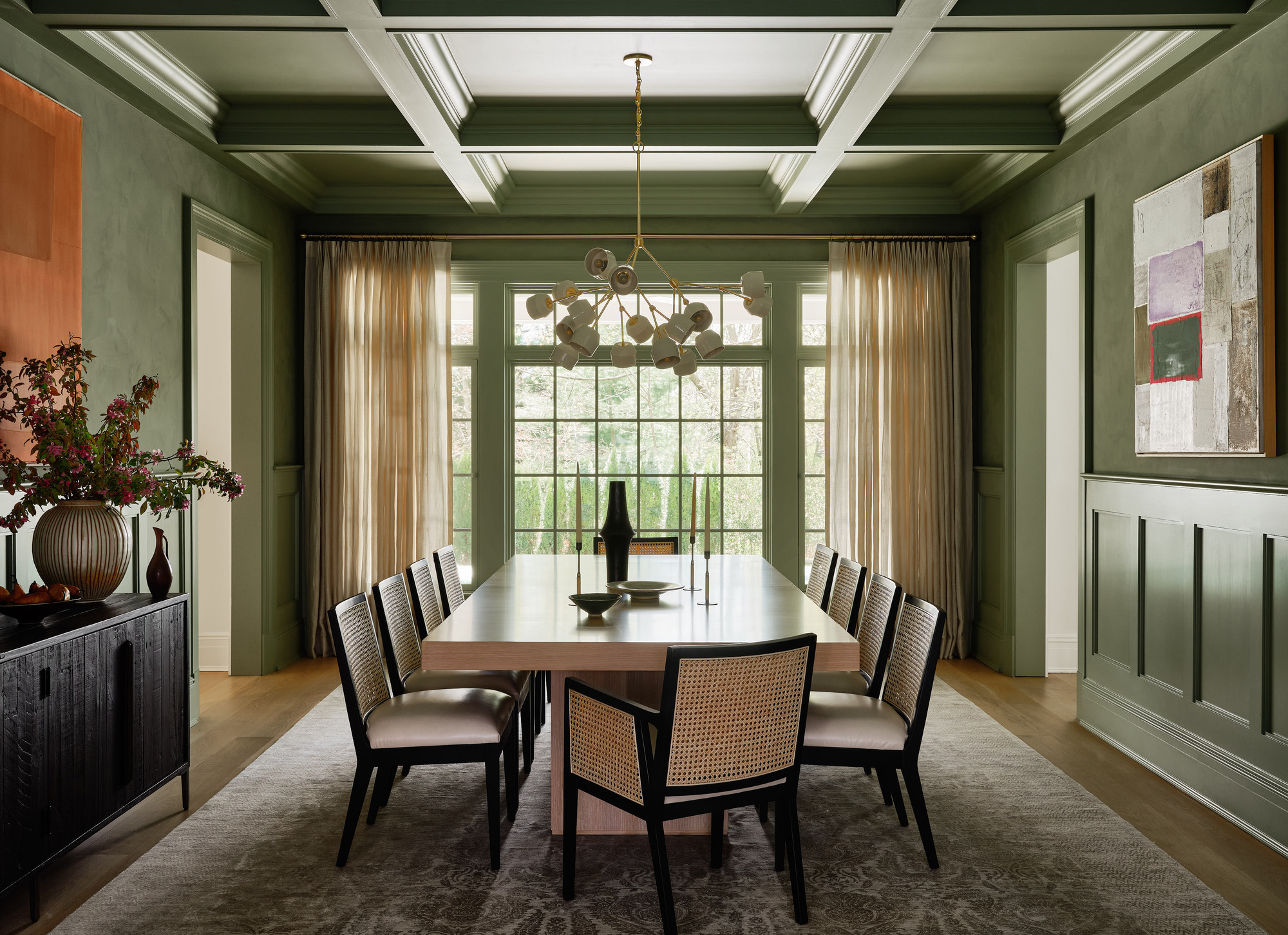 A green dining room painted with plaster walls, coffered ceiling, large dining table surrounded by chairs, a patterned rug, console table with vase of flowers and orange artwork