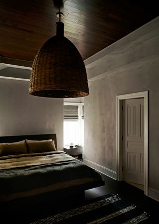 Dark bedroom with large woven lightshade and soft natural tones on the bedspread and grey plaster walls