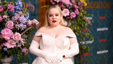 We love Nicola Coughlan's cocktail glasses. Here she is - a blonde woman wearing a white dress and white gloves with green rings, standing in front of a purple, blue, and pink flower display