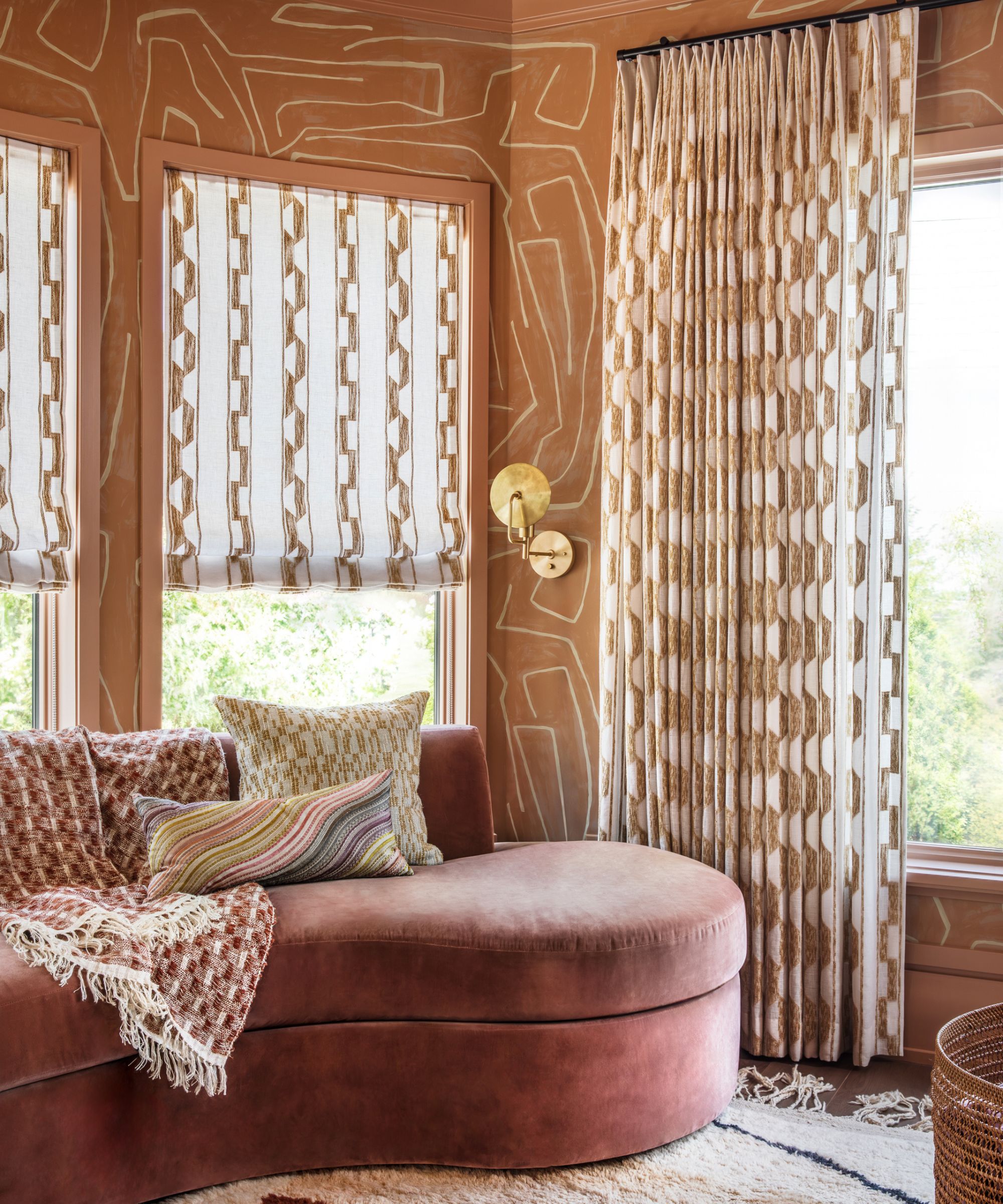 A decorative living room with blush pink wallpapered walls, large windows with neutral patterned window treatments, and a curved pink velvet sofa with patterned cushions.