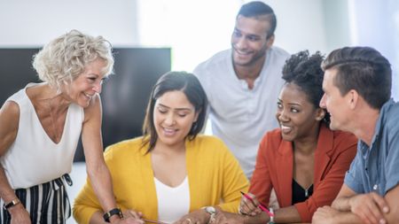 A diverse group of people are having a meeting in an office. They are sitting at a table with documents scattered on it, and having a meeting.