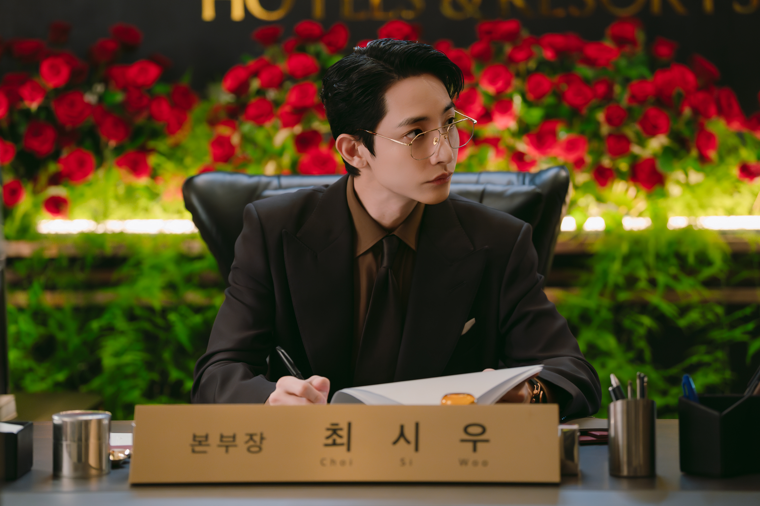 A man wearing reading glasses (Lee Soo-hyuk as Choi Si-woo) sits in behind a desk in an office full of red roses. A large nameplate in front of him reads &amp;quot;Choi Si Woo.&amp;quot; A still from the Netflix K-drama 'Boyfriend on Demand.'
