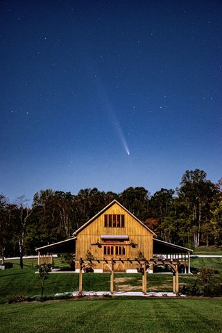 A light brown barn stands in a grassy field, with trees lining the horizon in the background. Above, in the dark blue night sky, a white comet streaks from lower right of center, with a tail jolting upward to the left.