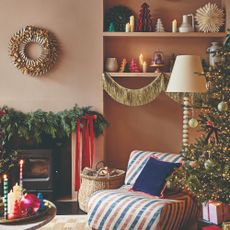 An earthy pink-painted living room decorated for Christmas with a lit-up Christmas tree, a garland-adorned fireplace and a gold tinsel garland decorating alcove shelving