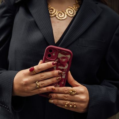 woman holding phone wearing gold rings and gold necklace with pinstripe black blazer and red phone case 