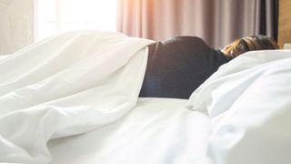 Woman lying in bed with covers over her, facing away from the camera, with curtains open and sunlight coming in