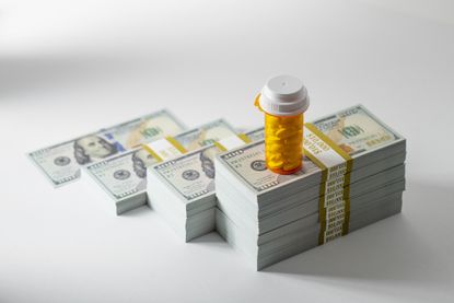 Prescription pill bottle with yellow tablets on growing stacks of US $100 bills, conceptual image representing pharmaceutical expenses, healthcare inflation and medication debt.