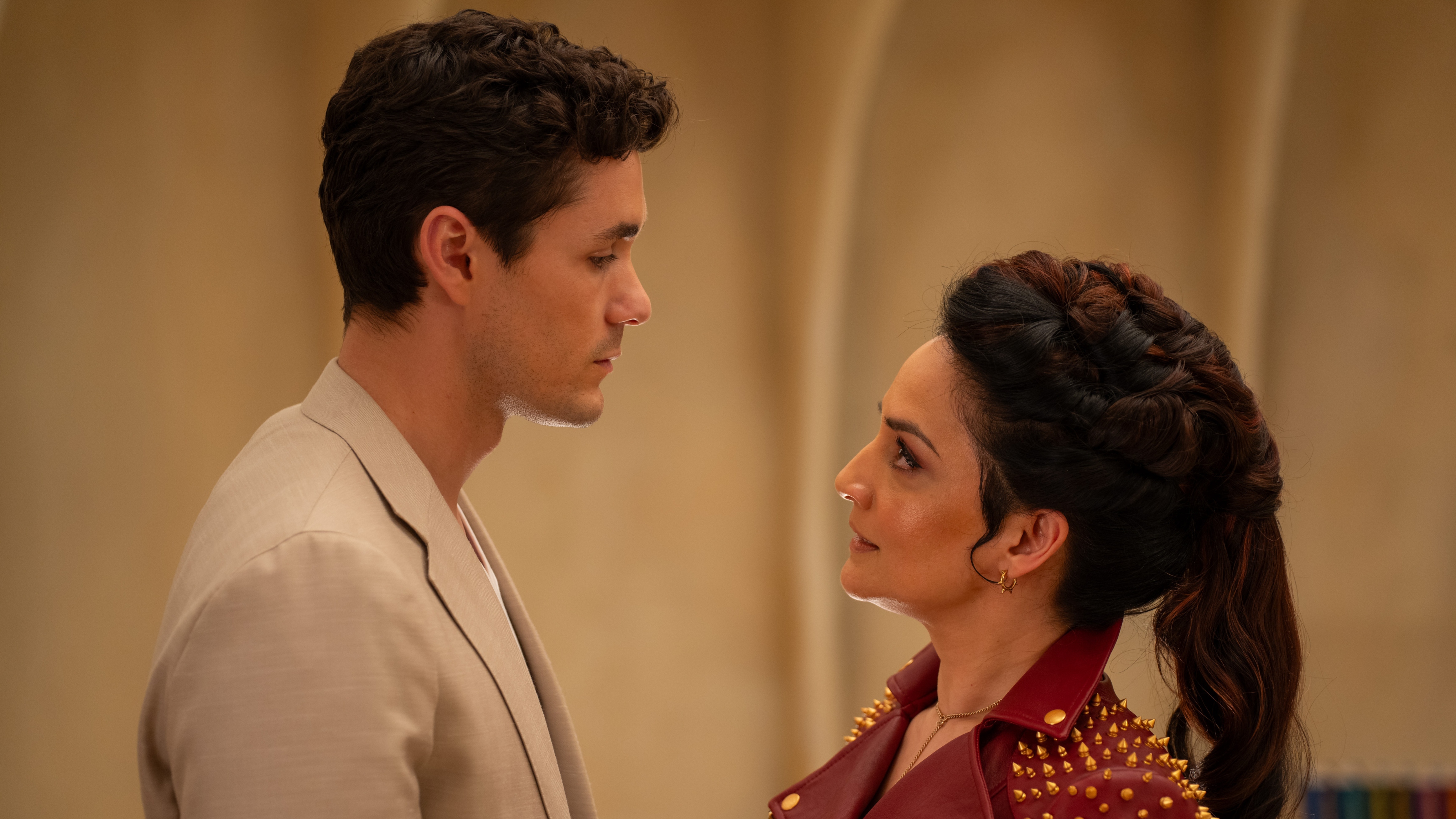 Conrad Clark (Jonah Hauer-King) stands opposite The Rani (Archie Panjabi) in a beige coloured room. He is wearing a stone-coloured suit and she is in a red leather jacket with small gold spikes all across the shoulders.