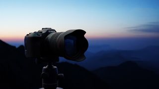 The silhouette of a Canon camera, mounted to a tripod, with mountainous blue hour backdrop