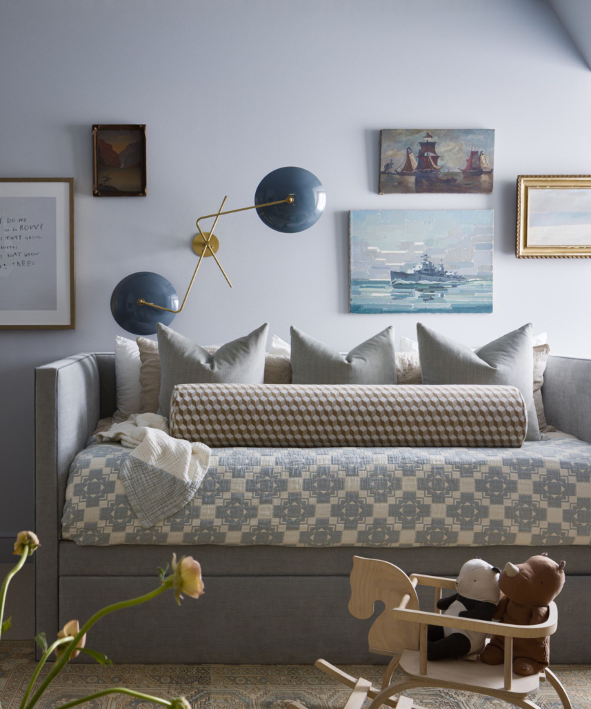 A child's room with light gray walls, a daybed with a light blue patterned quilt, a gallery wall, and a blue lamp.