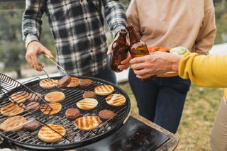 Three people standing at a BBQ, cheers-ing beers while flipping burgers.