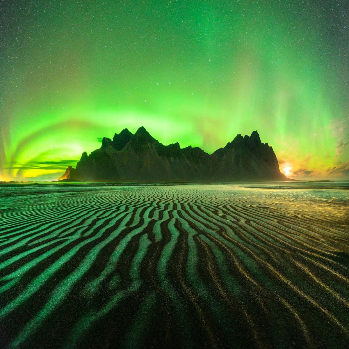 A mountain rises from a beach's wavy black sands, glowing in greens and pale yellows beneath the blazing aurora arched in the sky.