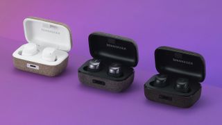 Tre stycken Sennheiser Momentum True Wireless 3 visas upp i sina laddningsfodral i färgerna vitt, mörkgrått och svart (öronsnäckorna). Visas upp mot en lila bakgrund.