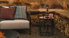 A sofa outdoors in a backyard with a firepit in the middle of the garden, beside the sofa is a side table with a stacked lamp on top and a red and white glass