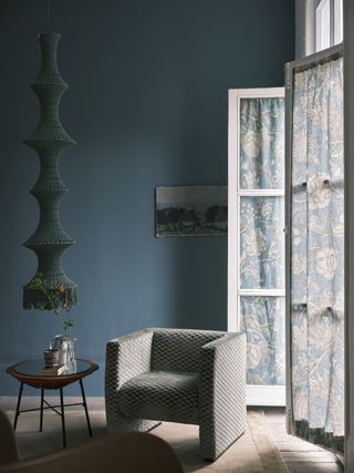 Image of a dark teal blue paint color in a living room with little light even though there are two French door windows open in the room. There is an accent chair and a small side table.