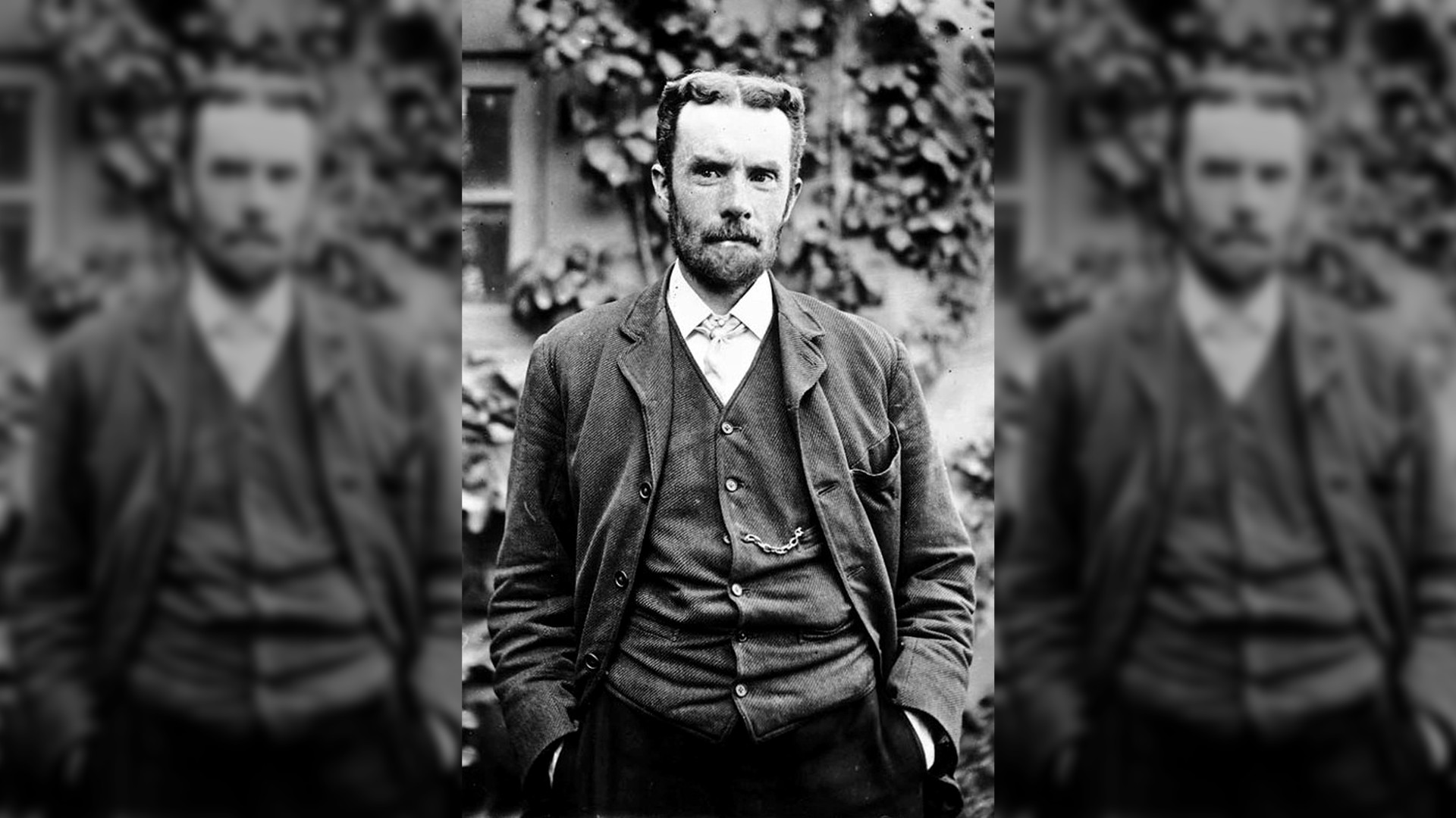 Black and white photo of Oliver Heaviside, English electrical engineer, mathematician, and physicist, standing in front of a tall hedge.