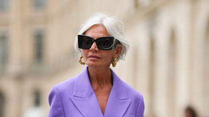 Grece Ghanem wears black sunglasses from Saint Laurent, a beige leather and gold braided earrings, a purple oversized blazer jacket, matching purple large suit pants, a dark brown shiny braided leather handbag, outside the COS show, on April 26, 2023 in Paris, France