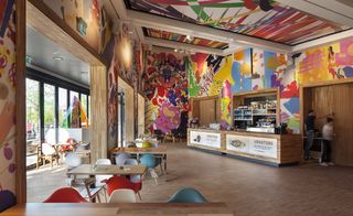 Inside view of the colourful dining area in the Eemcentrum building, wooden floor, wooden counter, people, wooden tables and colourful chairs, glass front entrance, colourful artwork on the walls and ceiling, white beams