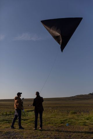 Daytime image, aerocene sculpture being flown by Tomás Saraceno and Frédéric Panaiotis, rope cord, grassy landscape, blue sky