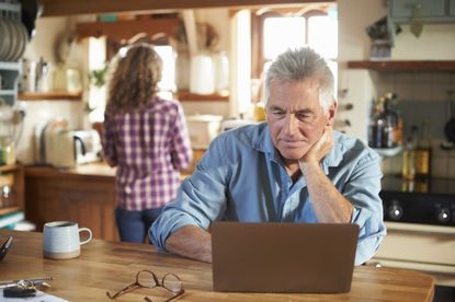 An older man looks at his laptop at the kitchen table. His female partner is seen in the background.