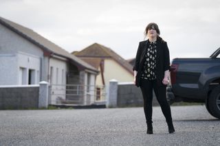 Tosh (Alison O'Donnell) stands in a driveway next to a pick-up truck. She looks a little hesitant. She is dressed up smarter than her usual police gear, in a black blazer and matching trousers, with a black and white floral top.