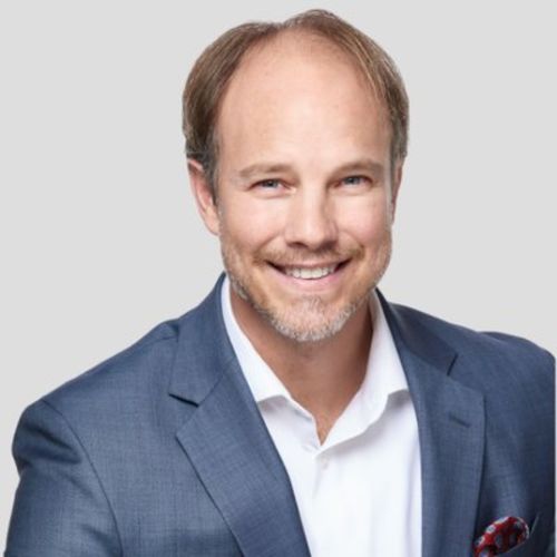 Tim Shurr is a white man wearing a medium blue suit and button down polo, he is pictured smiling at the camera on a gray background