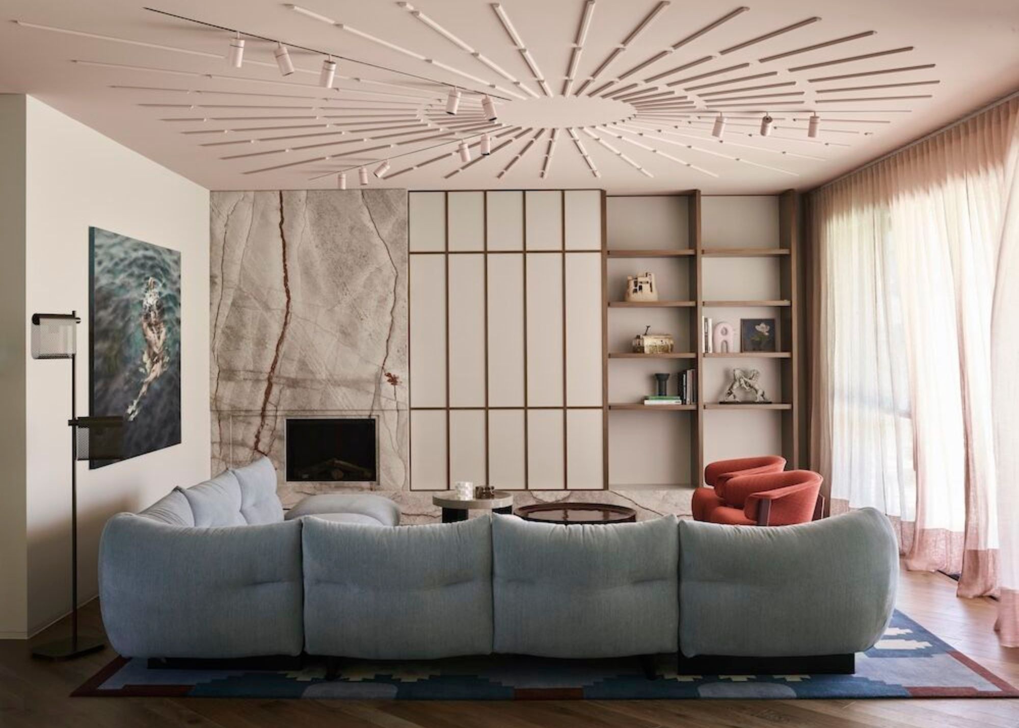 A pastel pink living room with marble fireplace wall, joinery, artwork on wall, starburst pattern on ceiling, light blue sofa, and sheer curtains with ruffles over the windows