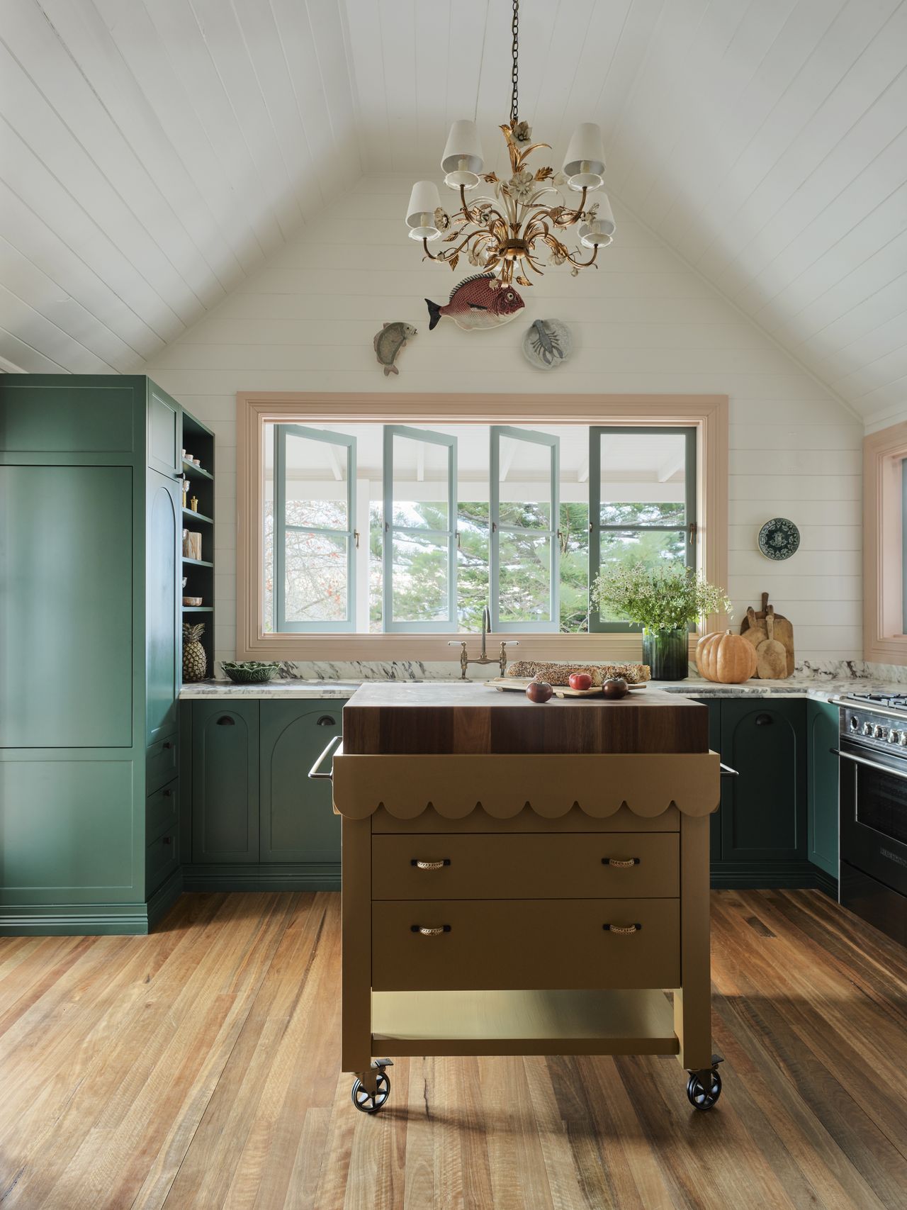 colorful kitchen with green cabinetry, marble trim, pink window frames, white timber paneled walls and ceiling, chandelier pendant light, and olive green kitchen island on wheels with scalloped edge detail