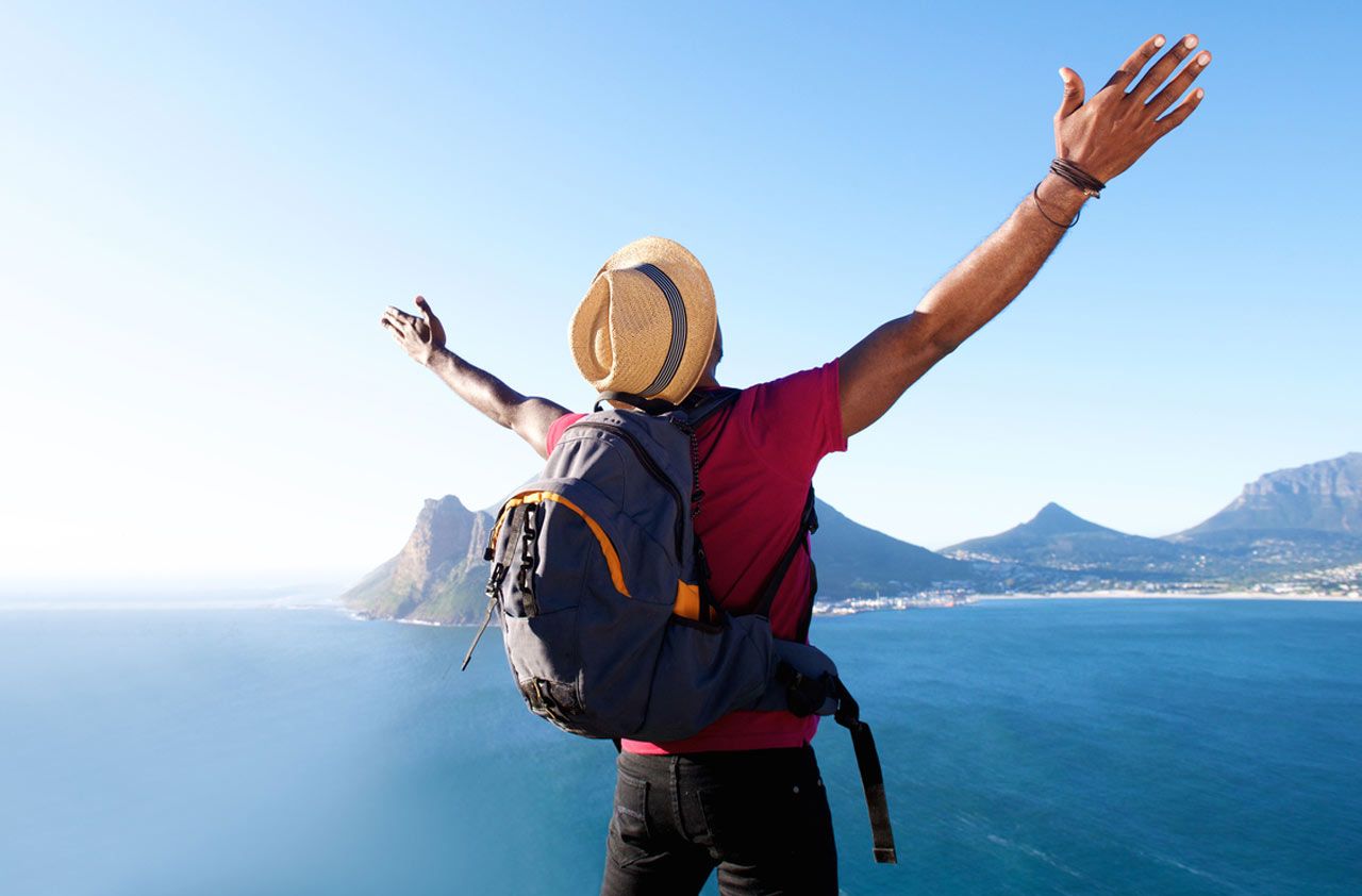 A hiker overlooks the sea with mountains in the distance and raises his arms in appreciation of the view.