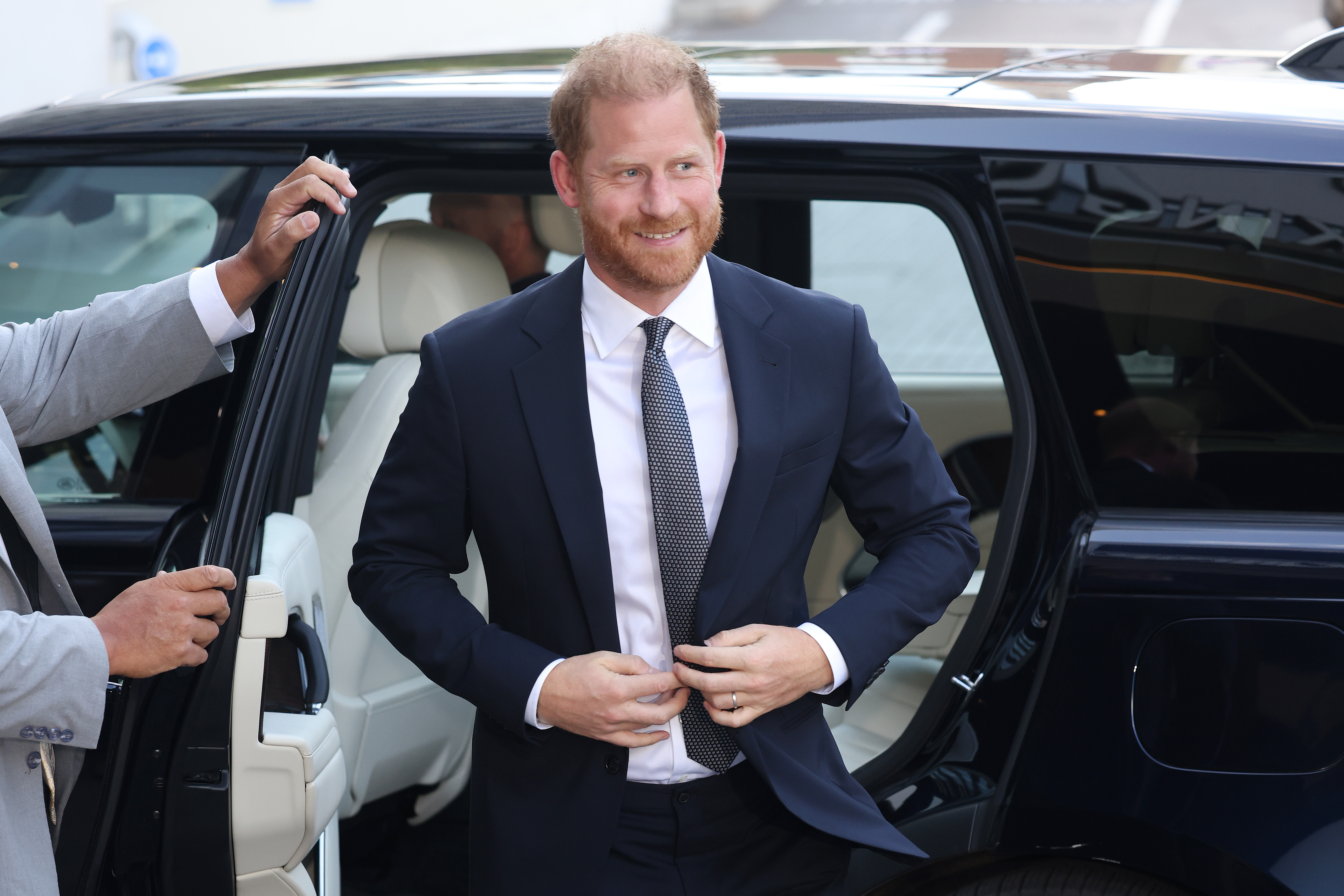 Prince Harry, wearing a navy blue suit and smiling, steps out of a black car