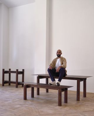 A portrait of designer Jerome Byron sitting on a table he designed and surrounded by more pieces of furniture from his collection, photographed at Carpenters Workshop Gallery