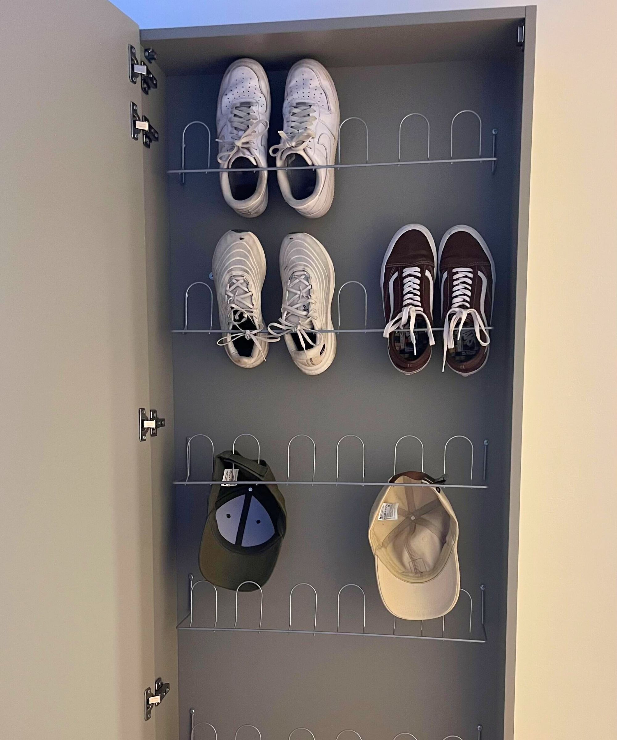 An open shoe cabinet with mounted shoe racks. Three pairs of shoes inside and two baseball caps.