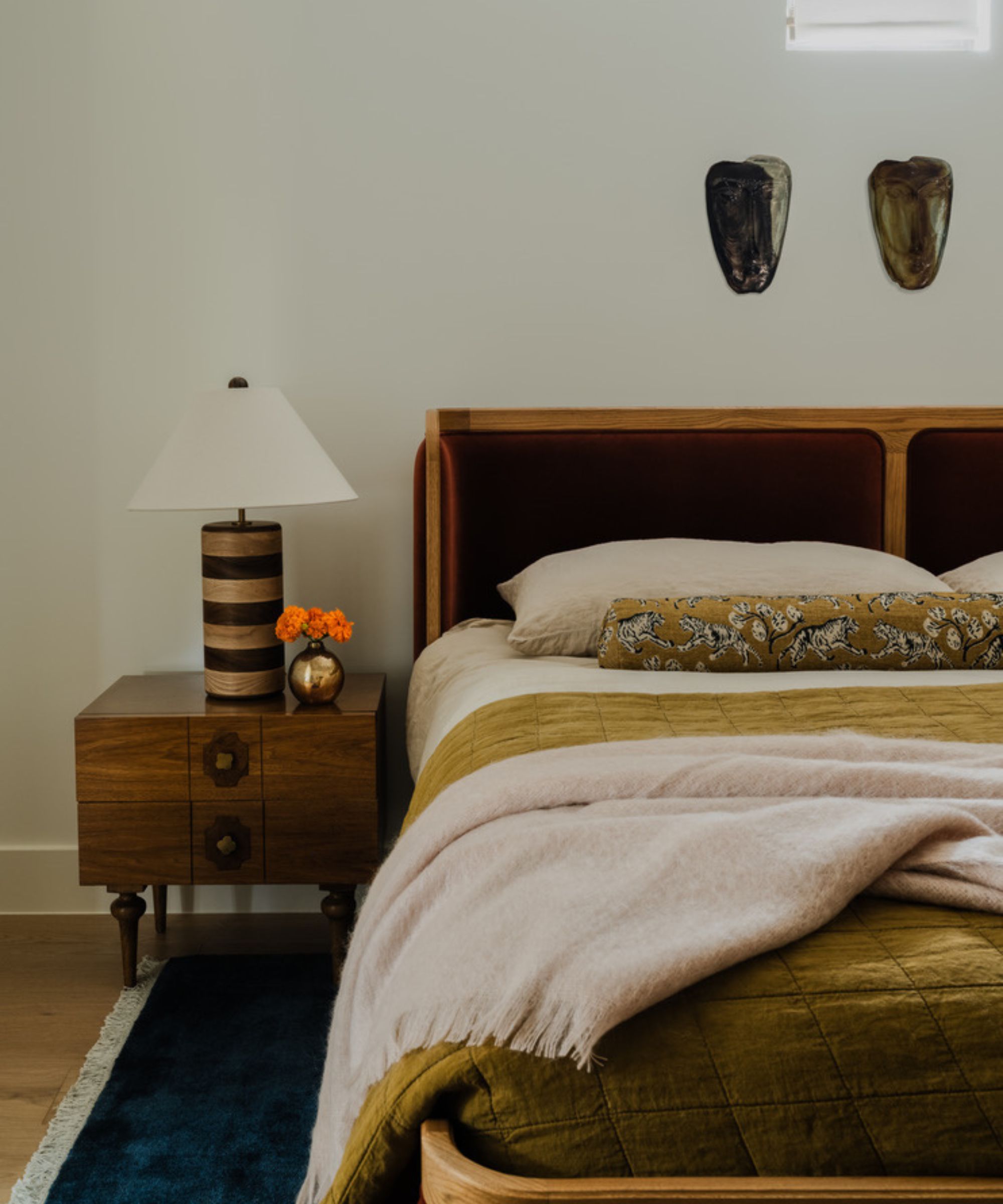 Fall bedroom with a dark red headboard, neutral bedding with an olive green quilt and bolster cushion, a wooden nightstand with a burl wood striped lamp and a dark blue rug.