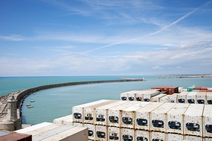 A large quanity of stacked shipping containers juxtaposed with the sweeping curve of a marina wall reaching out to the sea.