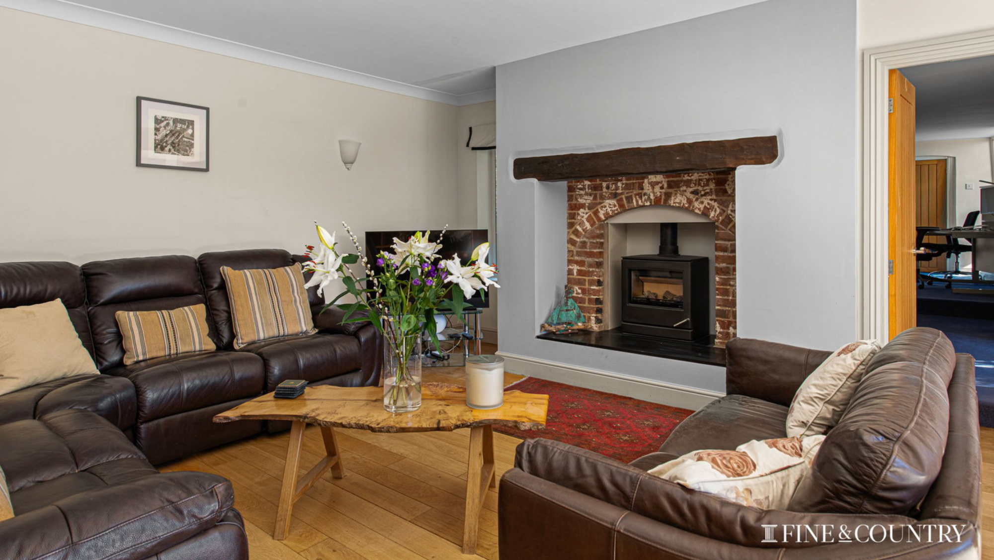 Living room with a wood floor, brick fireplace and wood burning stove. There is a wooden coffee table with a vase of flowers and brown settees either side of the room