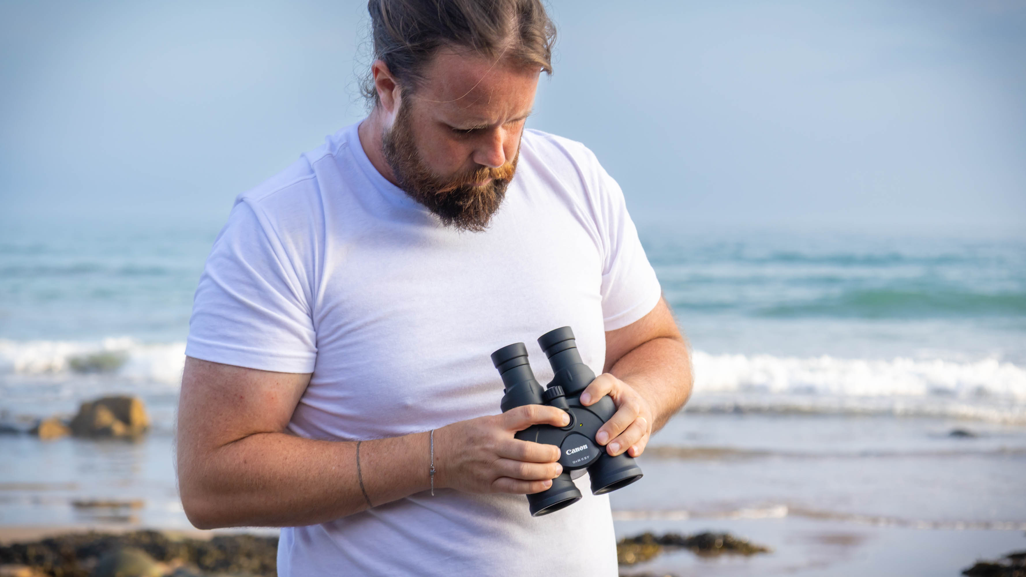 A male holding the Canon 12x36 IS binoculars and pressing the IS button, with the sea behind them.