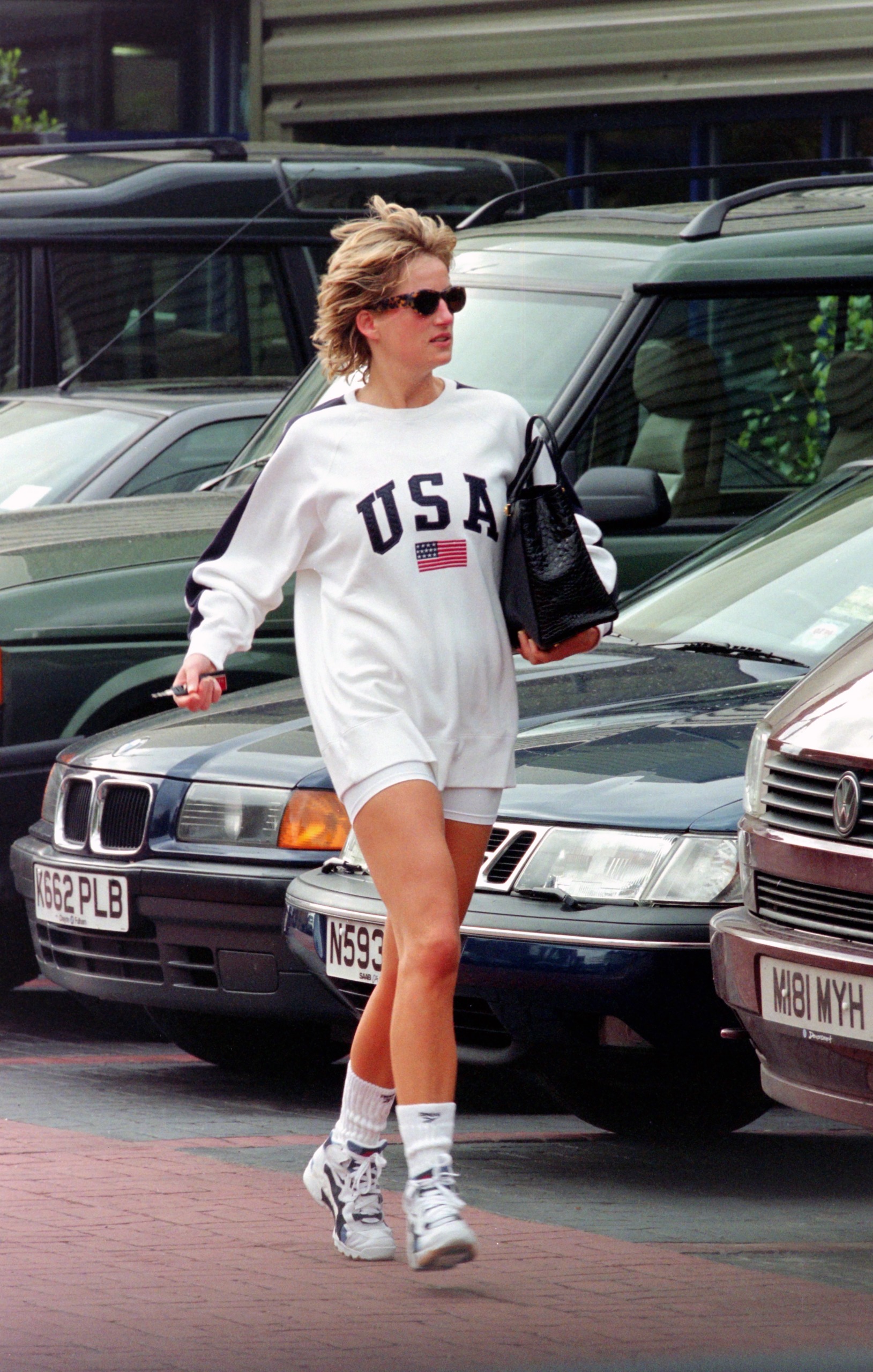 Princess Diana wearing a USA sweatshirt and bike shorts walking in front of cars