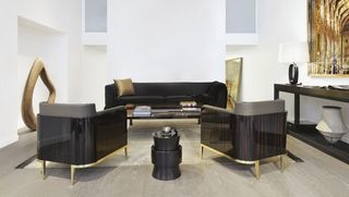 View of a space featuring white walls, wood flooring and various Christian Liaigre pieces of furniture including a black sofa with a bronze cushion, dark brown and gold armchairs and a black side table