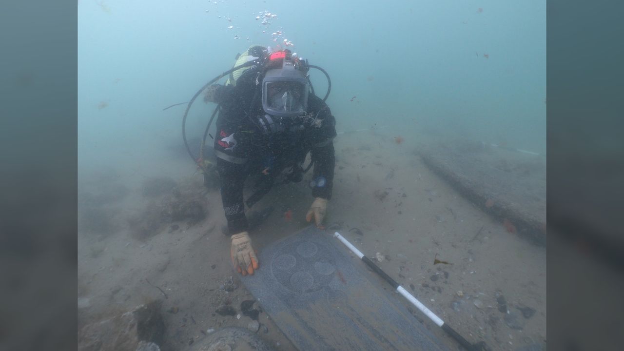 A scuba diver underwater showing off a gravestone decorated with Christian designs that was found on the seabed.