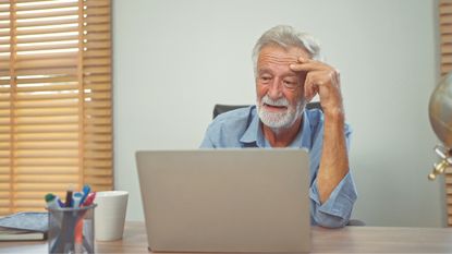 A worried older man uses a laptop at his kitchen table.