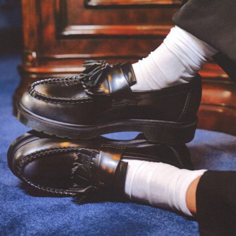 Photo of Dr. Martens Adrian Loafers in black with white socks, on blue carpet and desk behind. 