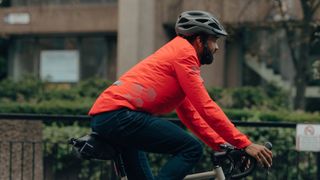 A man riding a bike in a helmet wearing the dhb flashlight orange waterproof jacket