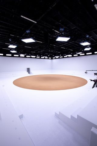 A studio space with a black ceiling, white wall, white floor and seating centred around a circular sand runway.